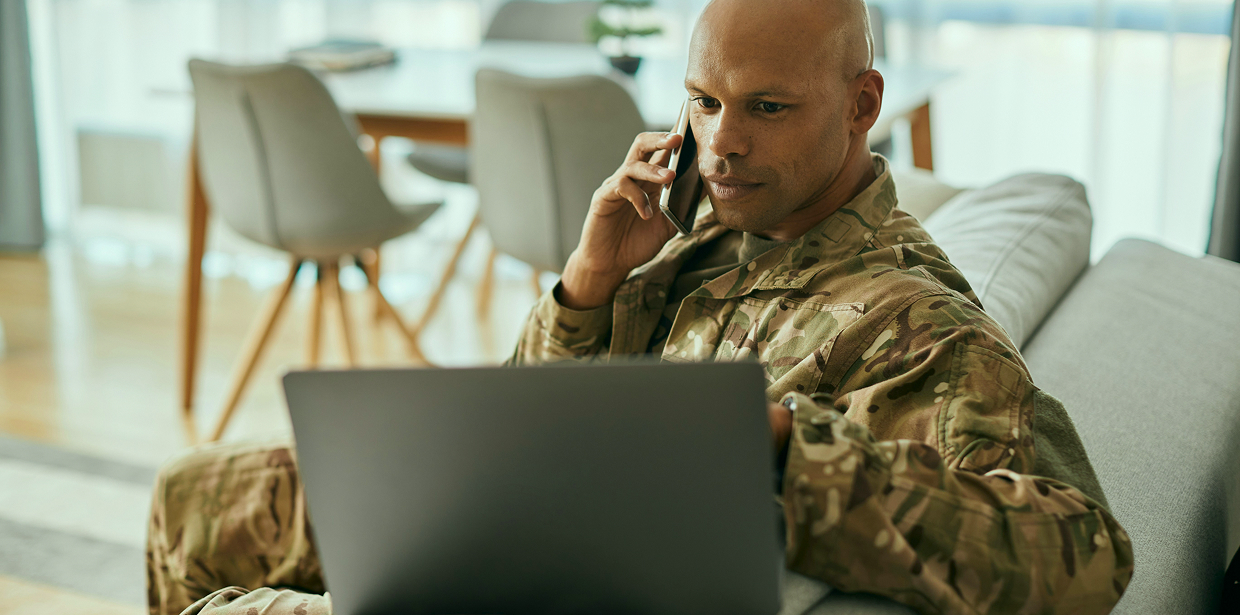 A service member in uniform sitting on a couch, talking on the phone while looking at a laptop