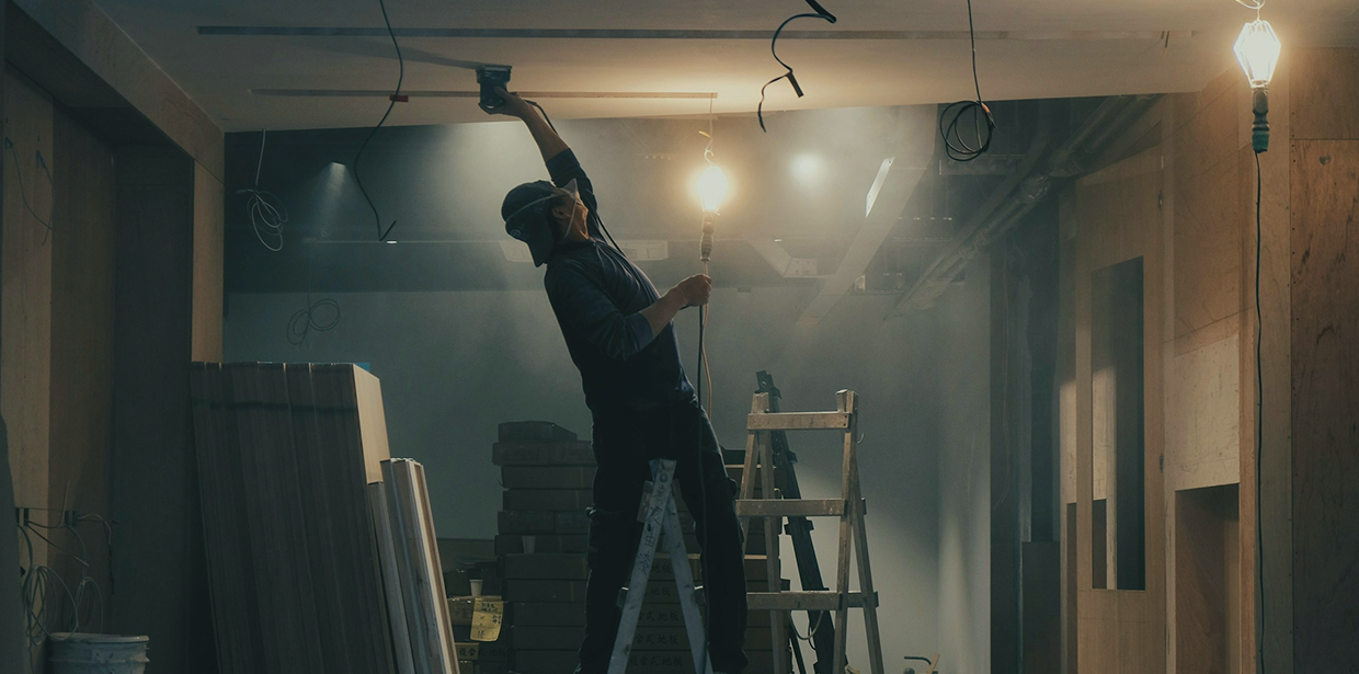 A construction worker on a ladder doing overhead work under job-site lights