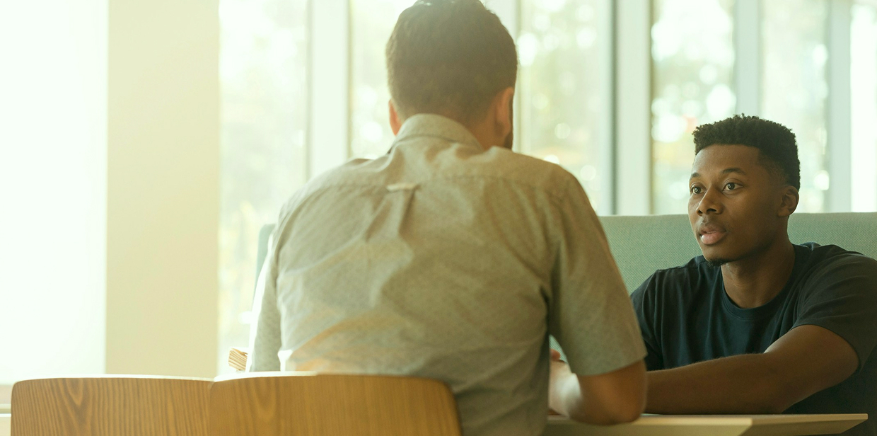 Two people speaking across a table in a sunlit room